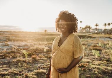 A smiling pregnant woman, wearing a yellow maxi dress, cradles her bump on the beach as the sun sets.