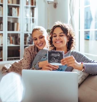 A happy female couple cuddle on the couch at home while showing an ultrasound of their baby on a video call via a laptop.