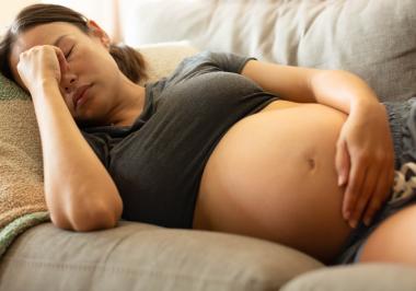 A tired pregnant woman lies on the couch with her eyes shut, resting one hand on her bump and the other on her forehead.