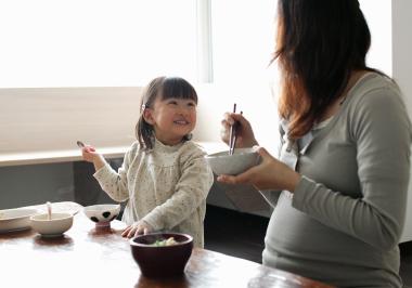 A little girl sitting at the dining table smiles at her pregnant mum, who is eating with chopsticks.