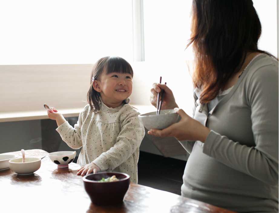 A little girl sitting at the dining table smiles at her pregnant mum, who is eating with chopsticks.