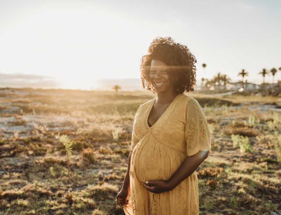 A smiling pregnant woman, wearing a yellow maxi dress, cradles her bump on the beach as the sun sets.