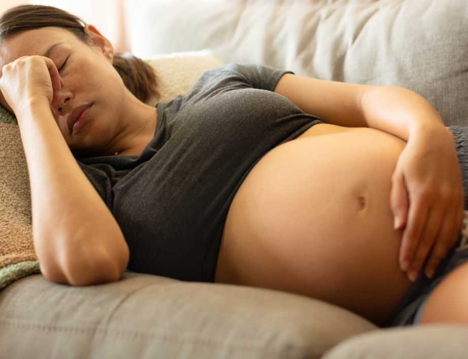 A tired pregnant woman lies on the couch with her eyes shut, resting one hand on her bump and the other on her forehead.