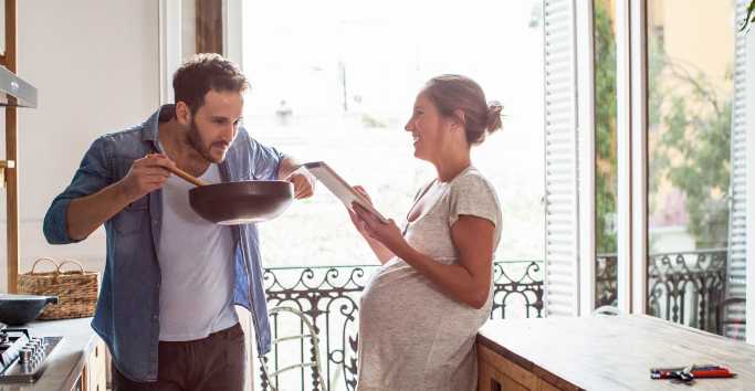 A pregnant woman holding a tablet laughs at her husband, tasting the meal he has cooked in a wok.